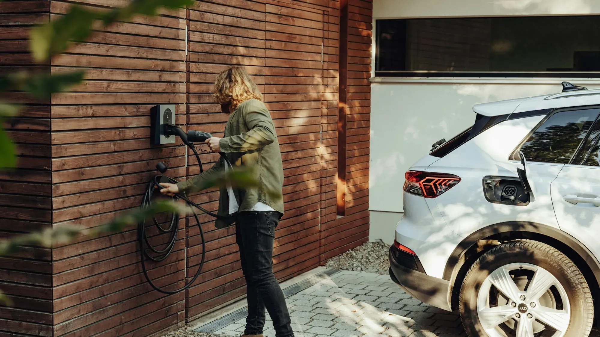 Persona conectando un carro eléctrico blanco a un cargador de pared en la entrada de una casa